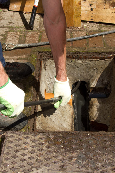 person using a plumber's snake to clear a drain with gloves on concrete surface demonstrating plumbing techniques and repair services for two different issues