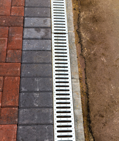 paved surface with red and gray bricks next to a drainage channel featuring bars and slots for water flow this design includes two distinct colors and improves outdoor drainage systems